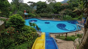 Piscina con tobogán, rodeada de abundante vegetación y montañas al fondo.