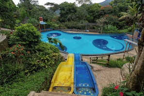 A swimming pool with a slide, surrounded by lush greenery and a mountain backdrop.