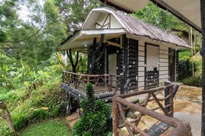 Een rustieke chalet met een houten terras en een rieten dak, omringd door weelderige begroeiing.