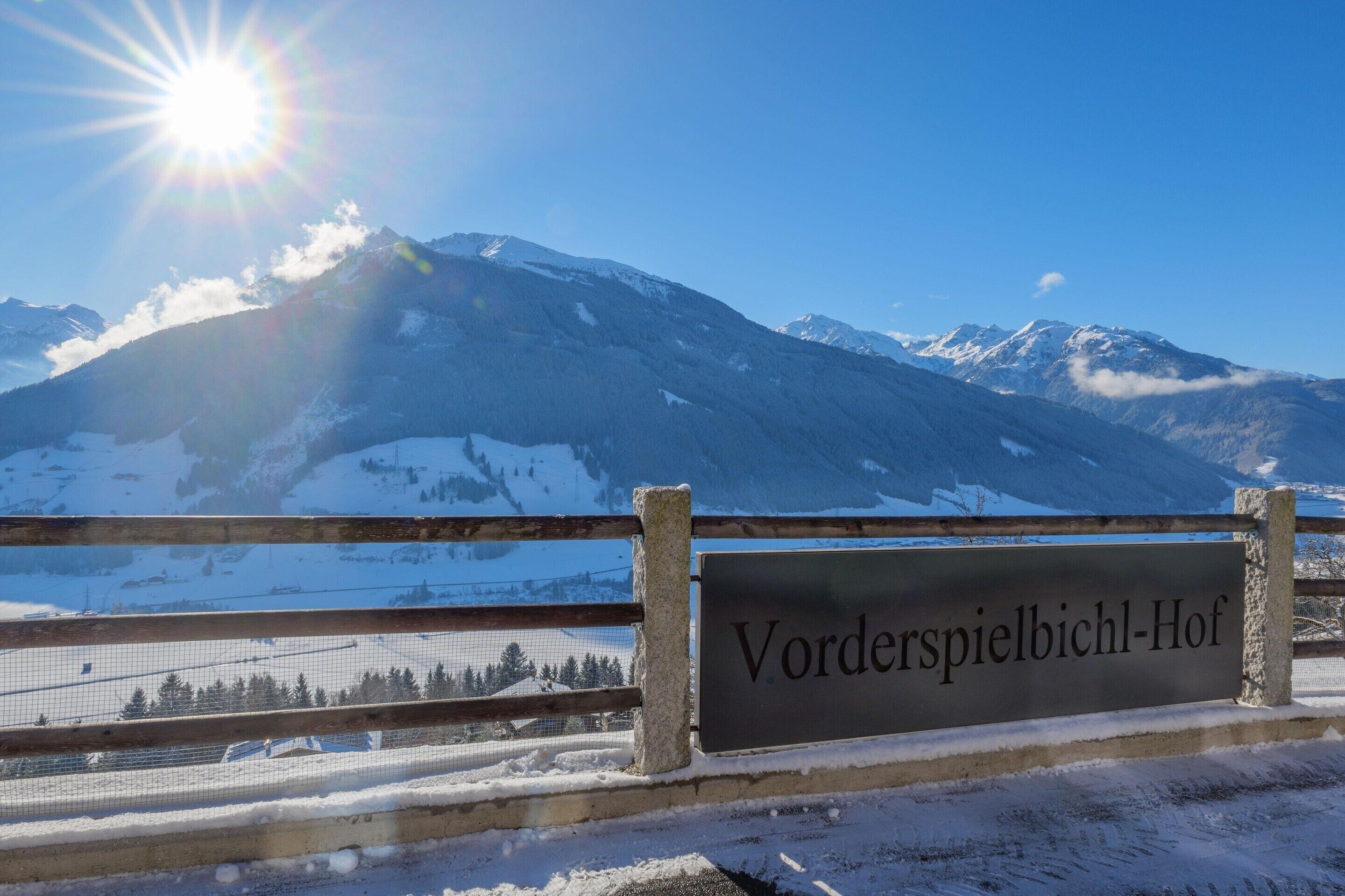 A sign with 'Vorderspielbichl-Hof' text, a wooden fence, snow-covered mountains, and a clear sky.