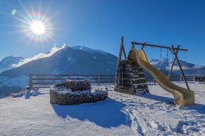 Ein verschneiter Außenbereich mit Holzzaun, Schaukel und einem mit Schnee bedeckten Holzstapel.
