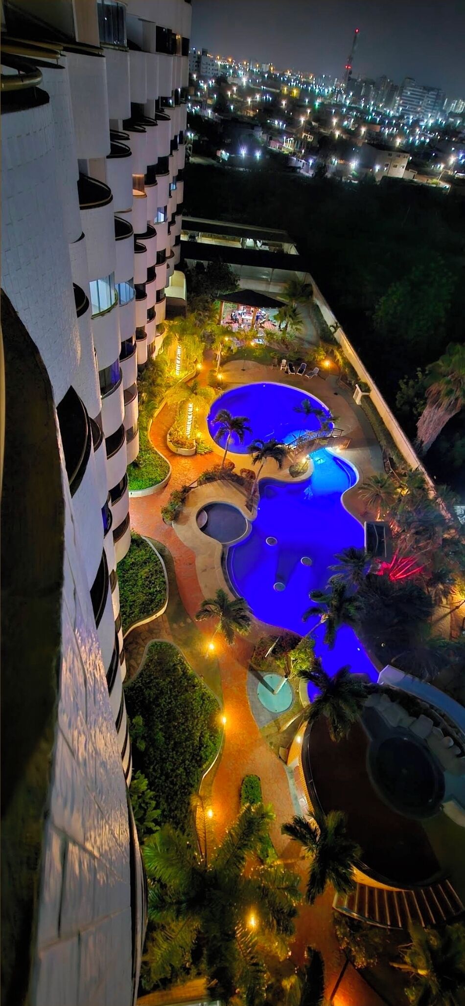 A rooftop pool area with multiple pools and palm trees, surrounded by a building with balconies.