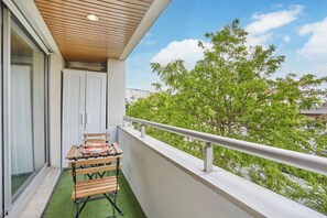 A balcony with a table set for two, a white door, and a view of trees and buildings. - Charming Studio Bercy Village/accor Arena (Paris)