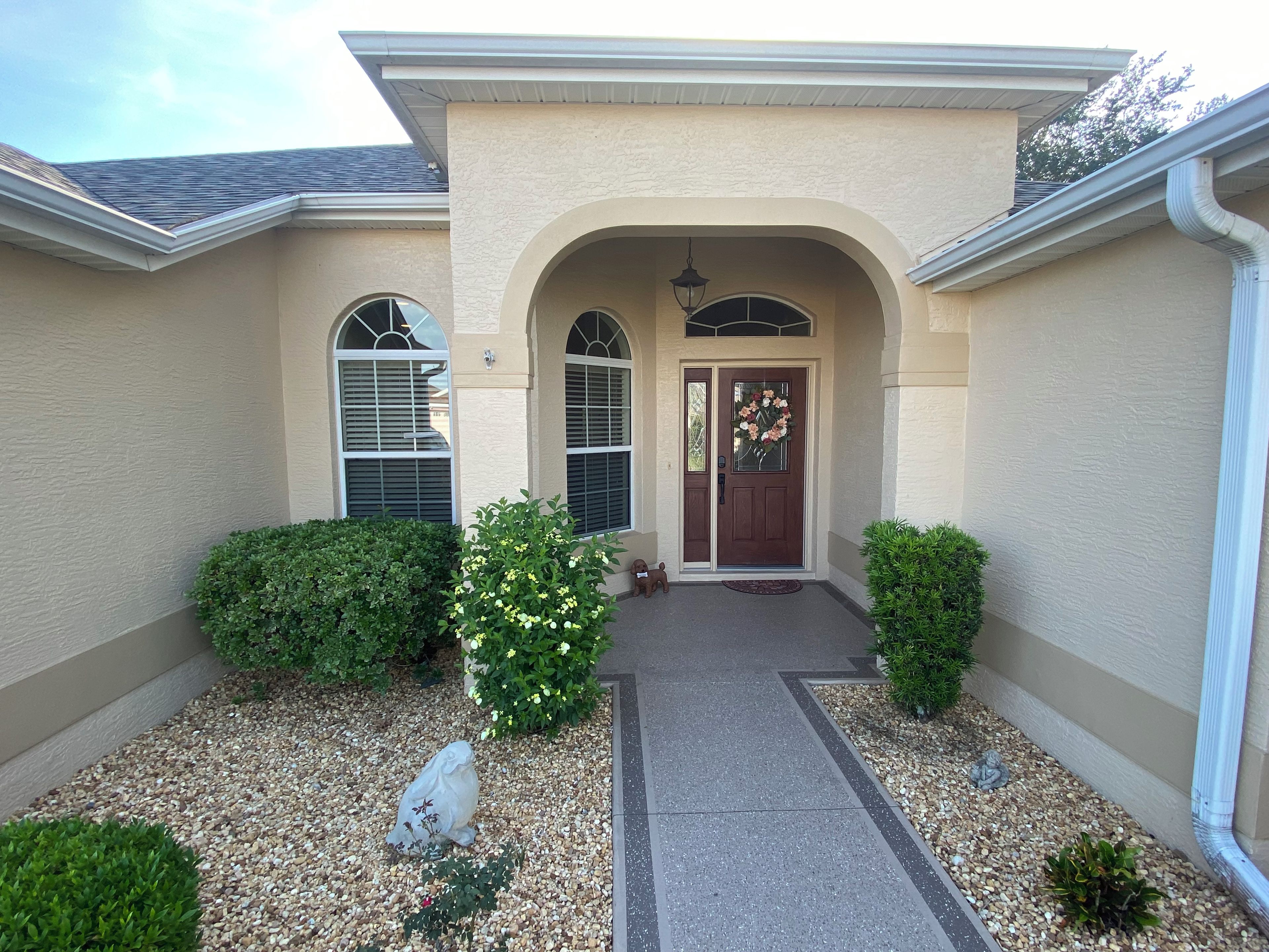 A residential building with a front entrance, two windows with shutters, a door with a flower wreath, and a small statue on the ground.