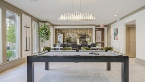 Une table de billard dans une pièce bien éclairée, agrémentée d’un lustre moderne, de fauteuils à revêtement vert et d’un coin bar avec un mur orné d’un motif hexagonal.
