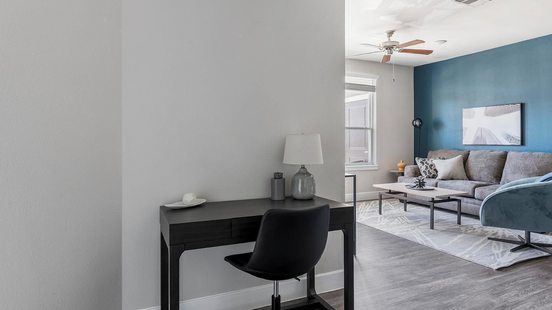 Uma sala de estar moderna com uma escrivaninha de madeira escura, uma cadeira preta, um sofá cinza e uma parede azul de destaque.
