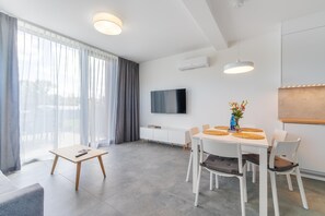 A modern living room with a dining area, large windows with curtains, a flat-screen TV, and a white air conditioning unit. - Coastal Holiday Home for 6 in Dziwnow (Dziwnów)