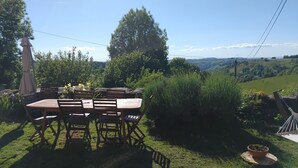 Ein Holztisch mit Stühlen draußen, mit Blick auf eine grüne Landschaft und Hügel im Hintergrund.