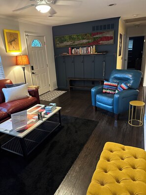 Ein Wohnzimmer mit Sofa, Sessel, Couchtisch und einem gelben Hocker. Eine Bücherreol steht voll mit Büchern und Dekoobjekten, eine Tür mit Buntglasfenster und ein Deckenventilator sind ebenfalls zu sehen.