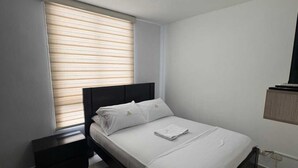 Ein Schlafzimmer mit Bett, Nachttisch, einem Fenster mit Jalousien und einer Wandleuchte.