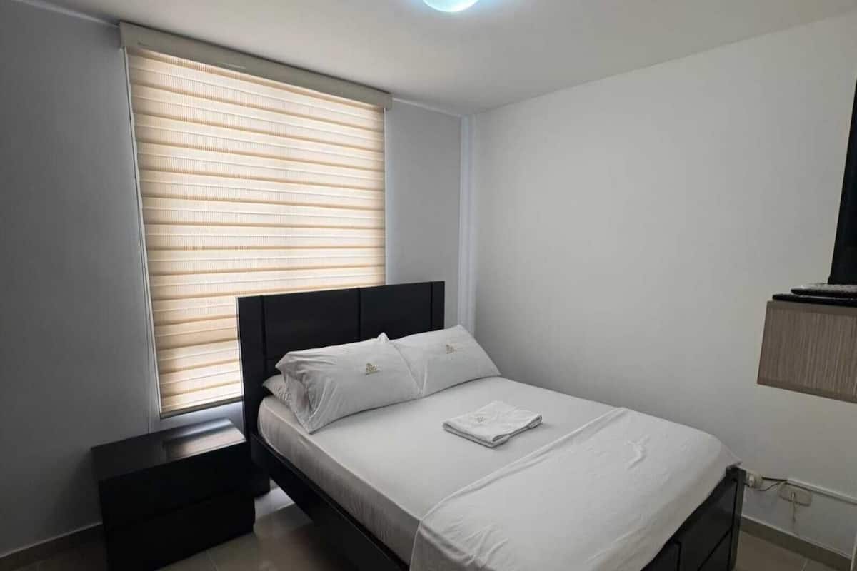 Une chambre à coucher comprenant un lit, une table de chevet, une fenêtre avec des stores et un luminaire fixé au mur.