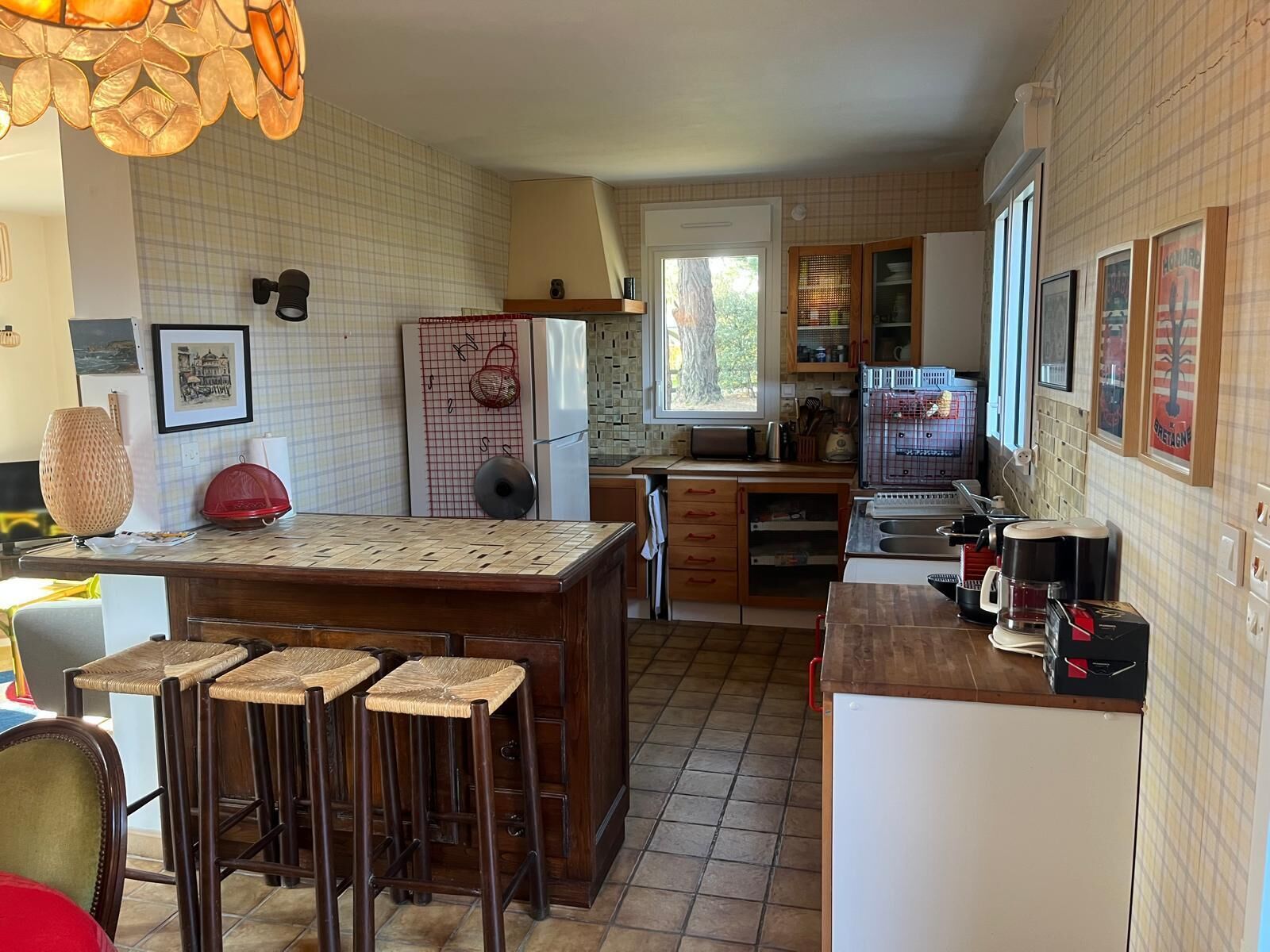 Cozinha com ilha central, banquetas, geladeira, máquina de café e janela com vista para árvores.