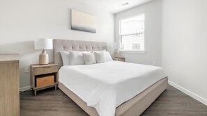 Een slaapkamer met een bed, nachtkastjes, een raam met jaloezieën en een kunstwerk aan de muur.