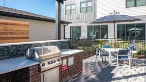 Een terras buiten met een barbecue, een tafel met stoelen en een parasol.