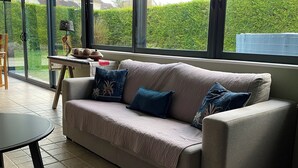 A modern living room with a beige sofa, a small table, and a view of a green garden through large windows. - Quiet apartment very close to Fontenay Abbey (Marmagne)