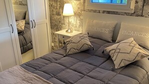 A bedroom with a bed, a nightstand with a lamp, a wardrobe, and a window with a view of greenery. - Quiet apartment very close to Fontenay Abbey (Marmagne)