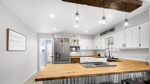 A modern kitchen with white cabinets, a wooden island, and hanging pendant lights. - The Cozy Escape (Los Lunas)