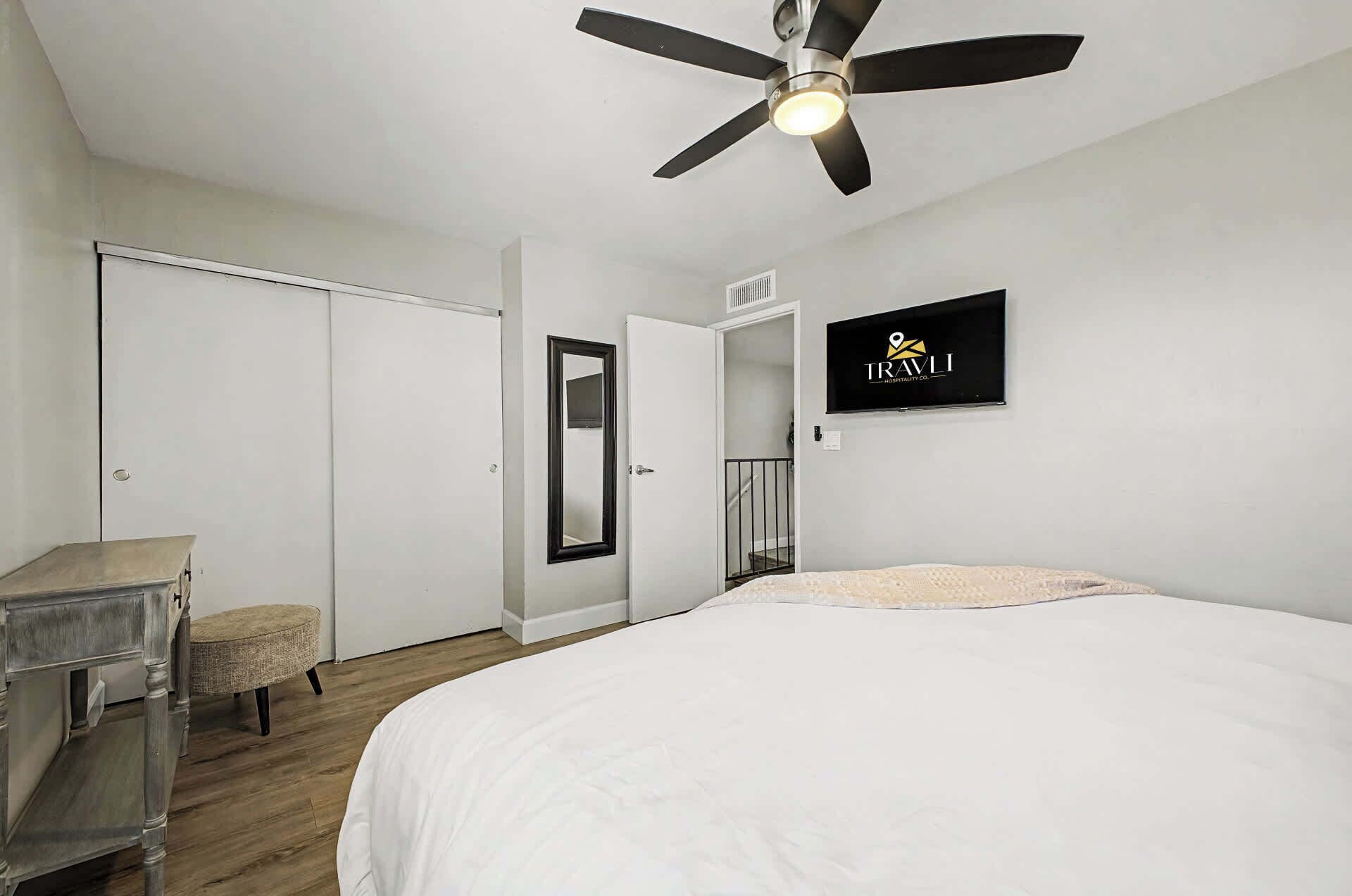 Een slaapkamer met een bed, plafondventilator, kledingkast, spiegel en een aan de muur bevestigde televisie.