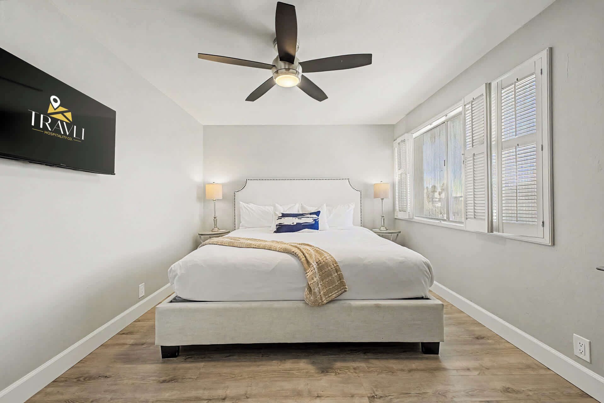 A bedroom with a bed, a ceiling fan, bedside lamps, a wall-mounted TV, and a window with blinds.