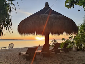 A beach scene with a thatched umbrella, lounge chairs, and a sunset over the water. - PILI BEACH RESORT AGMANIC (Santa Fe)