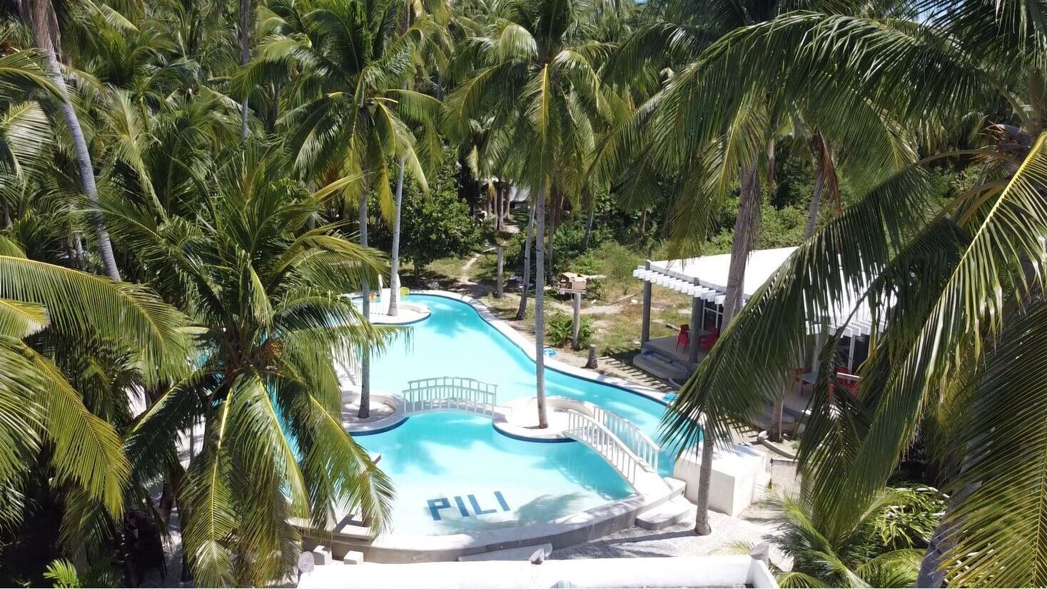 Piscina com a palavra 'PILI' na água, cercada por palmeiras e um edifício com telhado branco.
