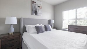 Habitación de hotel ordenada con una cama grande, dos mesitas de noche con lámparas, una ventana con persianas y un cuadro en la pared.