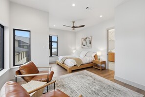 A modern bedroom with a bed, a chair, a bedside table, a ceiling fan, and a view of the outside.
