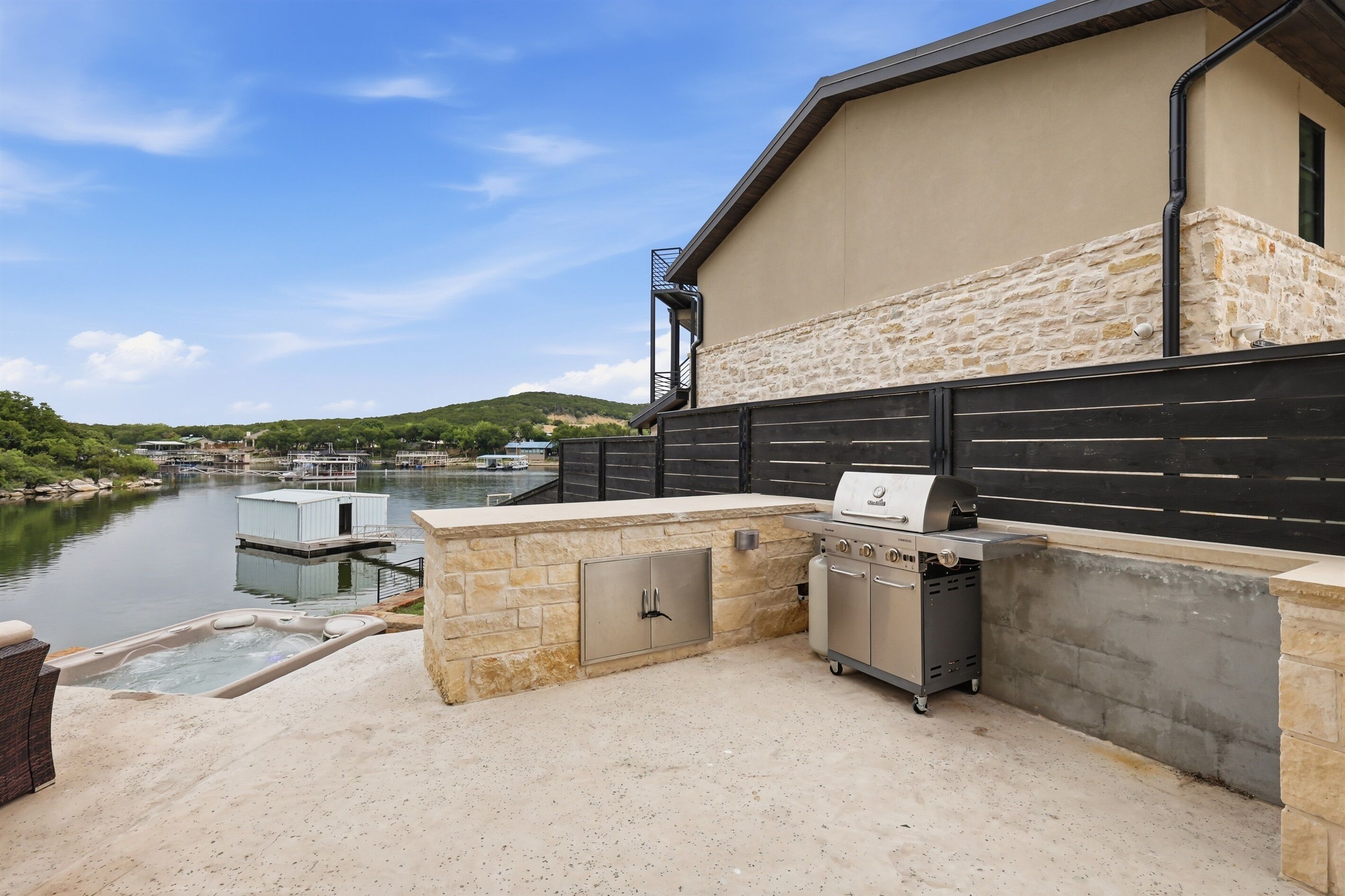 Een terras buiten met een barbecue, jacuzzi en stenen muren met uitzicht op een waterpartij.