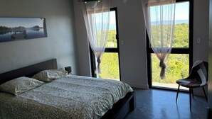 Quarto com cama, cadeira, quadro e janelas amplas com cortinas.