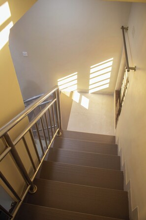 A staircase with metal railings and wooden steps, leading to a landing with a handrail and a wall-mounted light switch. - Monga's Apartments (Ndola)