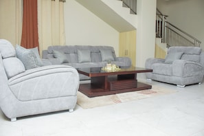 A living room with two grey armchairs, a grey sofa, a wooden coffee table, and a staircase in the background.