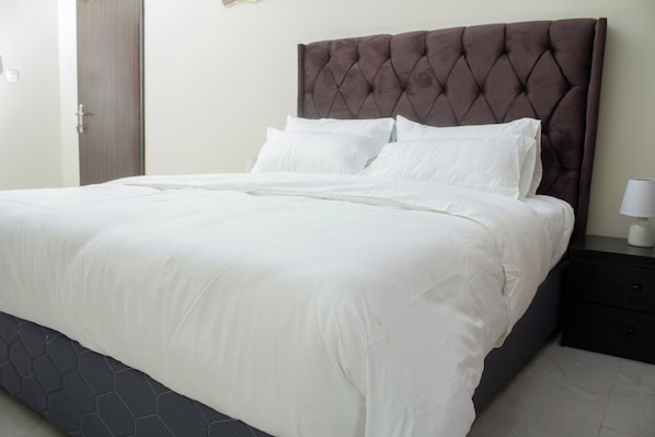 A neatly made bed with a tufted headboard, a bedside table with a lamp, and a door in the background.