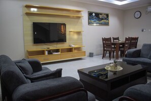 A living room with a flat-screen TV, wooden entertainment unit, dining table with chairs, and a sofa. - Monga's Apartments (Ndola)