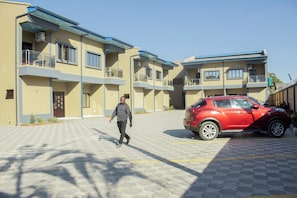 A building with multiple balconies, a red car parked in front, and a paved walkway.