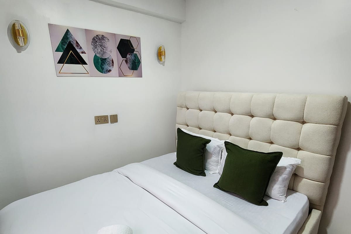 Una cama impecable con edredón blanco, almohadas verdes y blancas, y una toalla blanca enrollada.