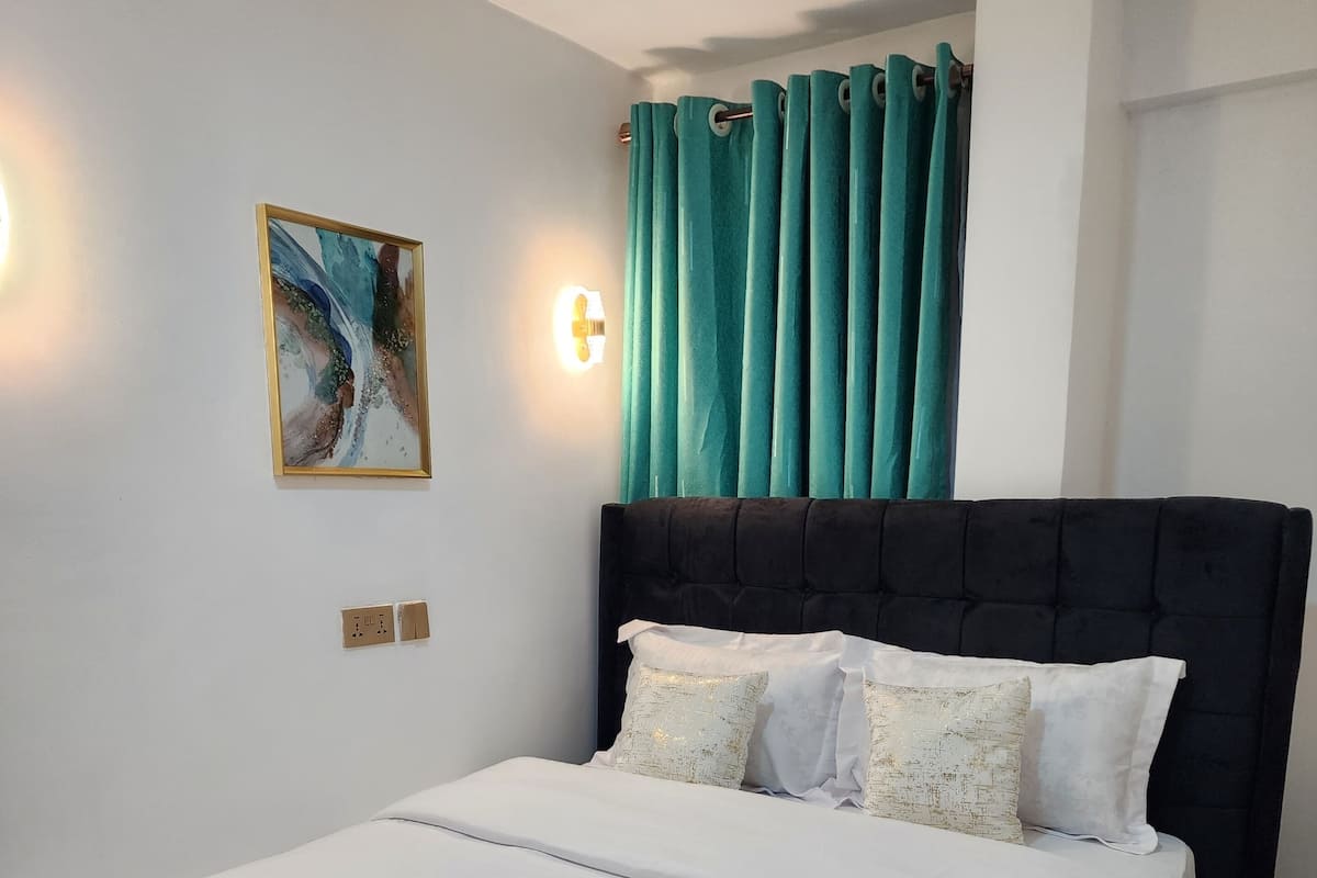 Una cama bien tendida con sábanas blancas y dos toallas enrolladas, un cabecero tapizado negro, cortinas azul verdoso y una lámpara fijada a la pared, junto con un cuadro enmarcado.