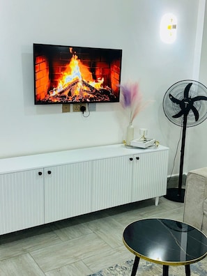 A living room with a TV mounted on the wall, a white cabinet, a black round table, and a standing fan. - BRIGIT LUXURY APARTMENT  (Kisumu)