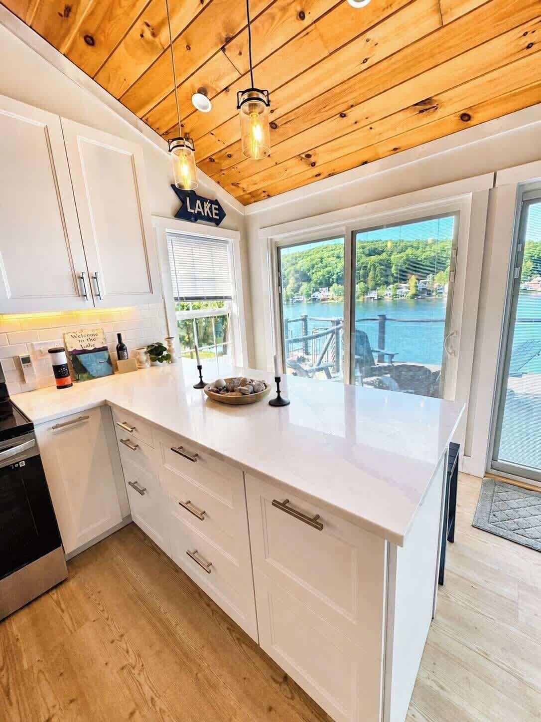 A kitchen with white cabinets, a wooden ceiling, and a view of a lake.