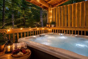 A hot tub in a wooden deck with string lights, surrounded by trees. - Cozy 3BR Cabin w/ Hot Tub, Pool Table & Arcade (Sevierville)