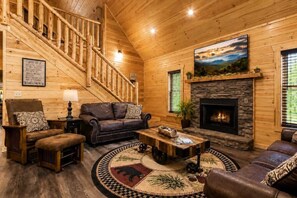 A log cabin interior with a fireplace, leather furniture, a staircase, and a painting on the wall. - Cozy 3BR Cabin w/ Hot Tub, Pool Table & Arcade (Sevierville)