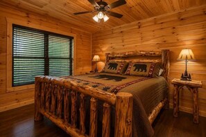 A wooden cabin bedroom with a bed, two bedside lamps, a ceiling fan, and a window with blinds. - Cozy 3BR Cabin w/ Hot Tub, Pool Table & Arcade (Sevierville)