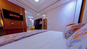 Een hotelkamer met een bed, een aan de muur bevestigde televisie waarop 'NETFLIX' staat, een stoel en een deur naar een andere kamer.
