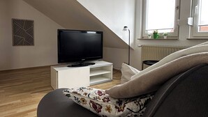 A modern living room with a flat-screen TV on a white stand, a window with blinds, and a radiator.