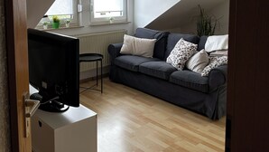 A modern living room with a dark grey sofa, a white side table, a flat-screen TV, a wooden floor, a window with blinds, a wall clock, and a radiator.
