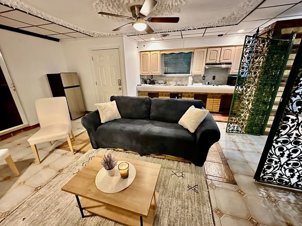 Un salon avec un canapé foncé, une table basse en bois, une chaise blanche et, au fond, une cuisine.