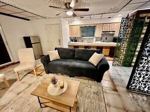 A living room with a dark sofa, a wooden coffee table, a white chair, and a kitchen in the background.