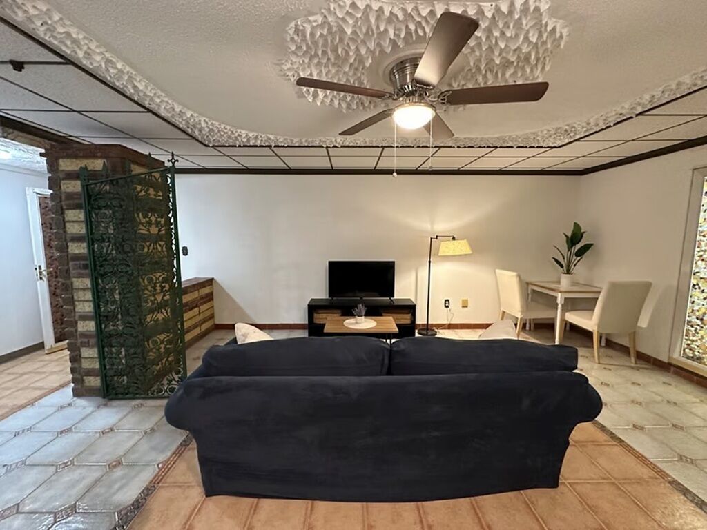 Un salon avec un ventilateur de plafond, un canapé noir, un téléviseur, une lampe et une petite table.
