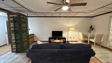 Un salon avec un ventilateur de plafond, un canapé noir, un téléviseur, une lampe et une petite table.