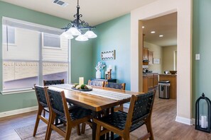 Un coin repas avec une table et des chaises en bois, un lustre, une fenêtre avec des stores et, au fond, une cuisine.