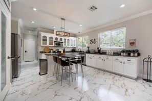 A spacious kitchen with marble flooring, a central island, bar stools, and a large window providing natural light. - The Mojave Tiki Mirage With Pool - North Las Vegas (North Las Vegas)